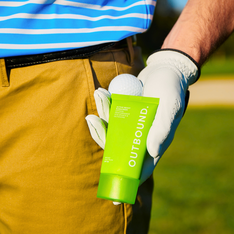 Golfer holding a green 'OUTBOUND' tube of sunscreen on a golf course